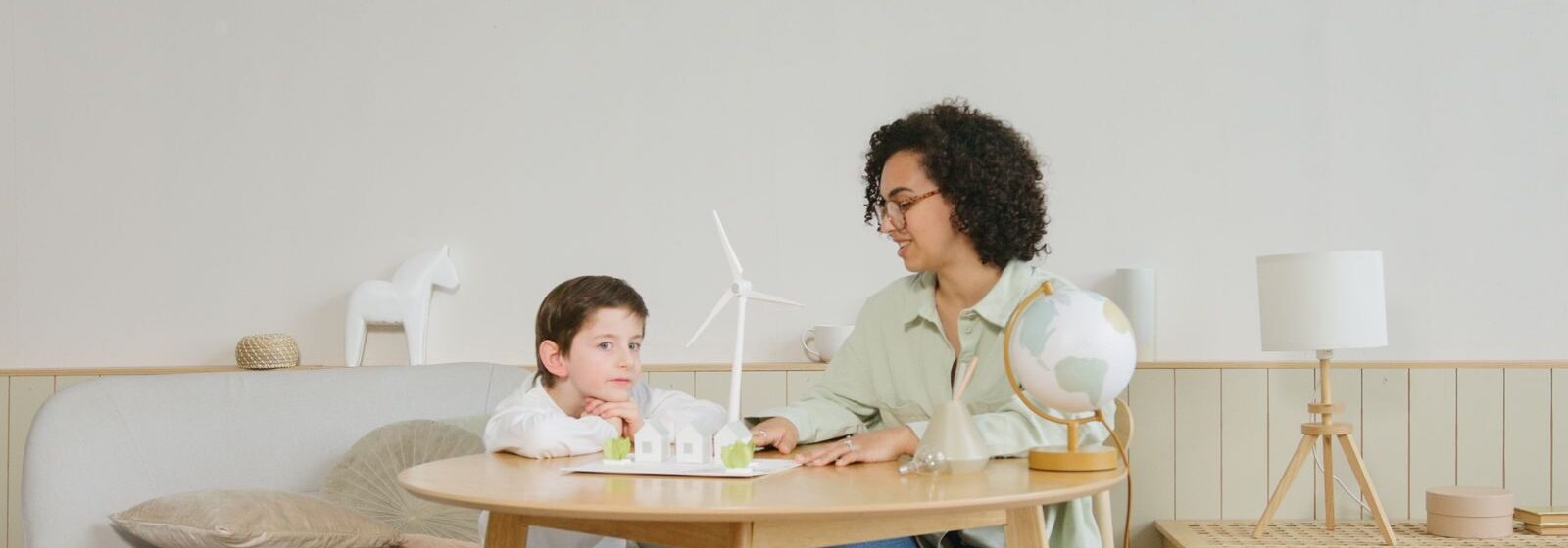A woman teaches a boy about science with a globe and windmill model in a cozy home setting.